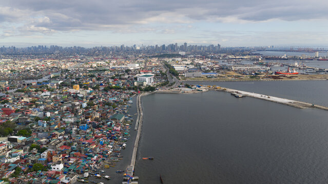 Aerial view of dense coastal housing and the busy harbor area with the distant city skyline under a cloudy sky in Navotas, Metro Manila, Philippines.