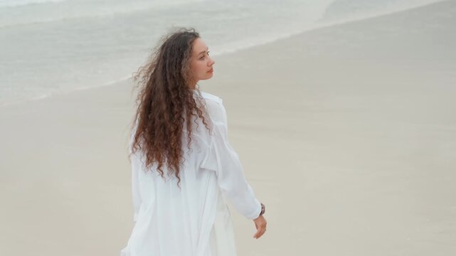 Woman walking along island sand, long curly hair flowing, white shirt billowing, gentle surf, barefoot steps leaving footprints, calm tide, empty horizon, travel mood, carefree pace