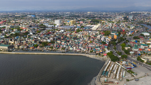 Aerial view of the dense residential area along the coastline with a breakwater and urban skyline under a hazy sky in Navotas, Metro Manila, Philippines.