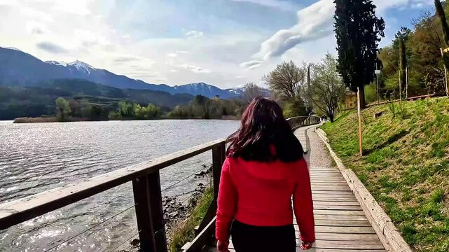 A woman, photographed from behind, walks on a walkway overlooking Lake Tublin in Trentino, Northern Italy. The woman has long black hair and wears a fuchsia down jacket