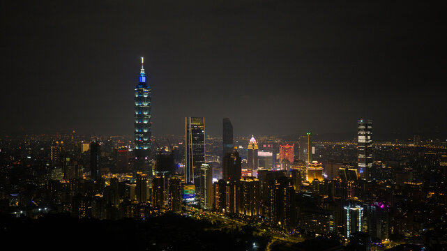 Aerial view of the Taipei 101 skyscraper and illuminated city skyline at night with glowing urban lights under a dark sky in Xicun Village, Taipei City, Taiwan.