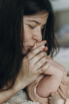 Happy Mothers day! Mother kissing newborn baby feet in bedroom. Love and family concept. Mom holding sweet little infant legs, close up. Adoption and maternity. Child health and care