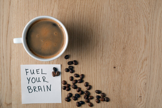 A cup of aromatic coffee and a note saying fuel your brain on a wooden surface with coffee beans. Concept of focus and productivity