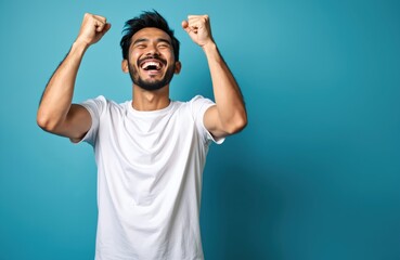 Naklejka premium Asian man with closed eyes smiles widely, raising clenched fists in celebration. Energetic young man wears white t-shirt, poses joyfully against blue background. His success and happiness are evident.