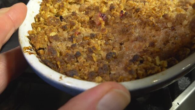Closeup of a large serving spoon breaking into a freshly baked, rhubarb crumble dessert in a dish, with crispy topping, and lifting out a spoonful.