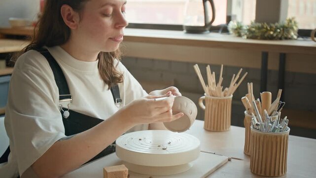 Woman using tool to apply pattern to clay cup, sitting at table with organizers with modeling tools in sunlit workshop