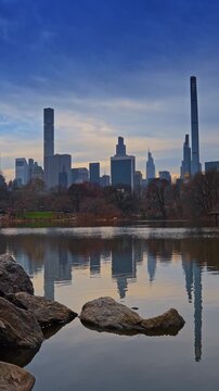 Boulders are at the waterfront of the pond in Central Park, New York, USA. Manhattan skyline against overcast sky in the backdrop. Vertical video.