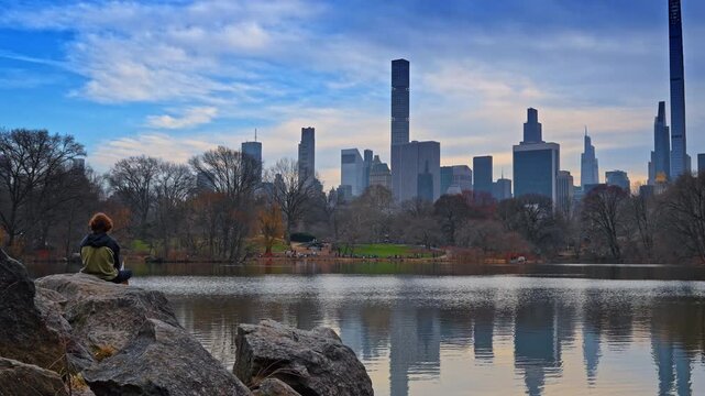 Rear view of a person sitting on the boulder near the pond in Central Park, New York, USA. Manhattan skyline against cloudy sky in the backdrop.