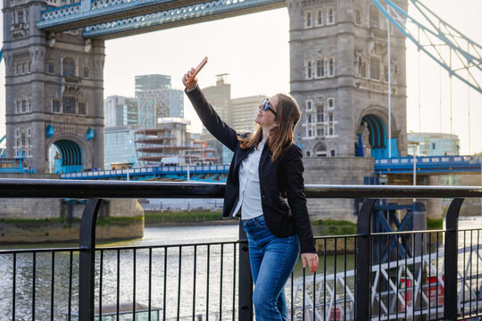 A happy tourist woman is taking selfie photos with her mobile phone in front of the Tower bridge in London, England