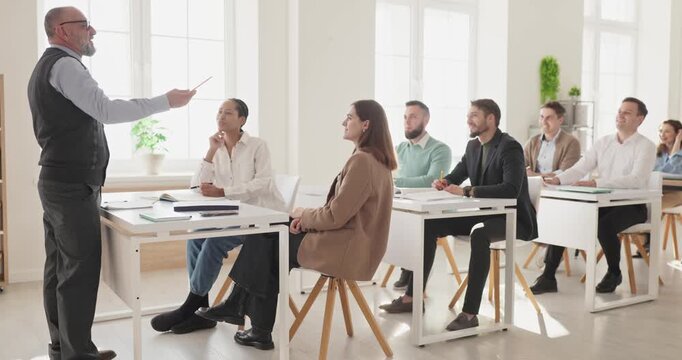 Teacher guides students in classroom training lecture. Diverse adults listen to a coach leading a business seminar, taking notes and joining discussion. Strong teamwork and leadership concept.