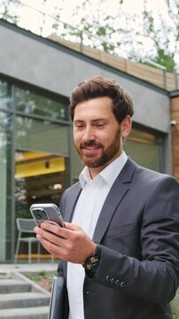 Confident businessman in a formal suit holding a laptop and using smartphone for online messaging or mobile work while walking in park area. Concept of digital communication, mobile applications