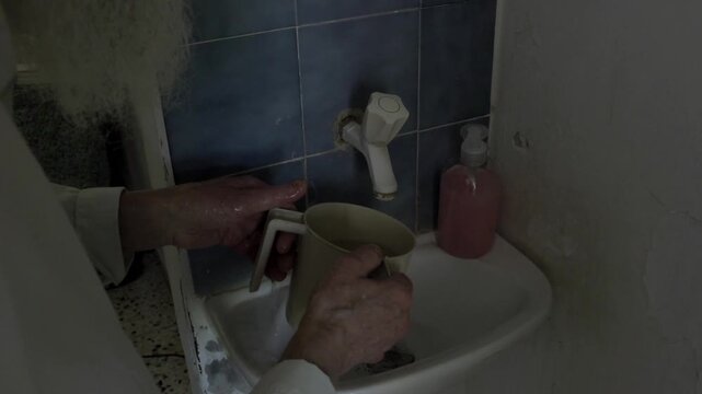 Old man with a long white beard performing a ritual washing of his hands in a small bathroom sink