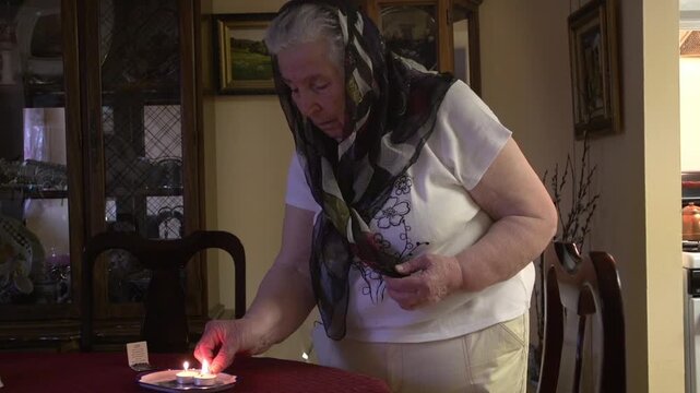 Senior Jewish woman with a head covering lighting candles for Shabbat and saying a prayer