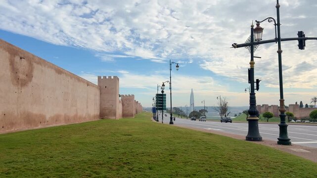 Ancient sandstone walls of the Chellah necropolis stretch along a modern road in RABAT, MOROCCO. Beyond the green lawn and busy traffic with cars and a stoplight.