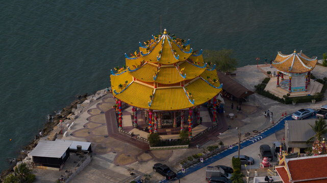 Aerial view of a traditional Chinese pavilion with a vibrant yellow tiered roof and ornate dragon pillars with people walking along the waterfront in Chonburi, Thailand.