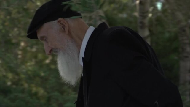 Pensive old jewish man with a long white beard and a kippah walking slowly in nature