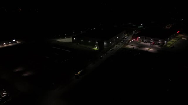 Aerial night flyover of large warehouse and industrial facility in United States with illuminated loading docks, trucks and surrounding infrastructure in dark commercial logistics area.