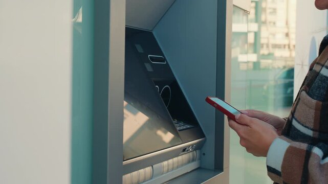 Close up of female hands holding smartphone and entering PIN code on ATM keypad. Cardless cash withdrawal, mobile banking, secure financial transaction and modern fintech technology.