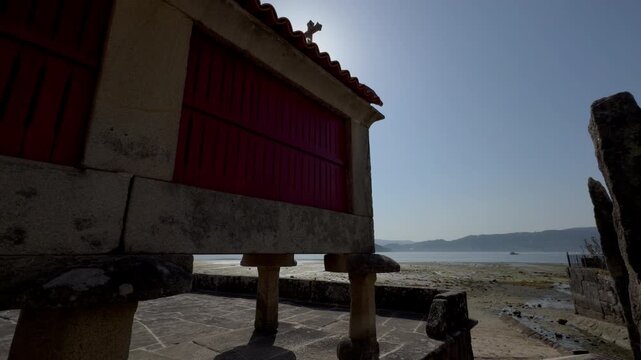 Backlit Galician Horreos on Estuary Shore with Sun Behind Cross