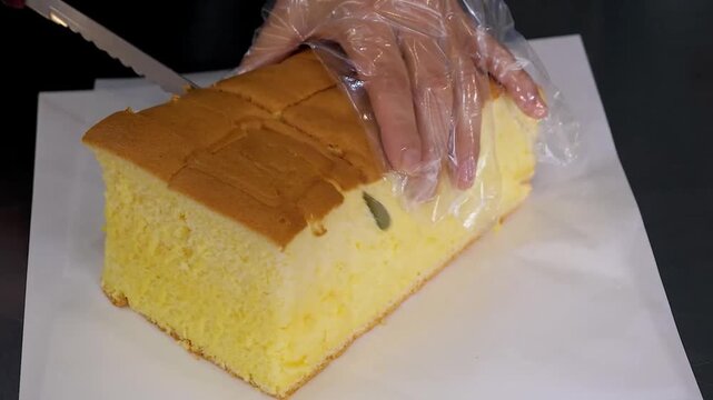 Close-up footage of a person wearing a clear plastic glove using a serrated knife to slice a large, freshly baked, soft, and jiggly golden-yellow Taiwanese Castella sponge cake on white parchment pape