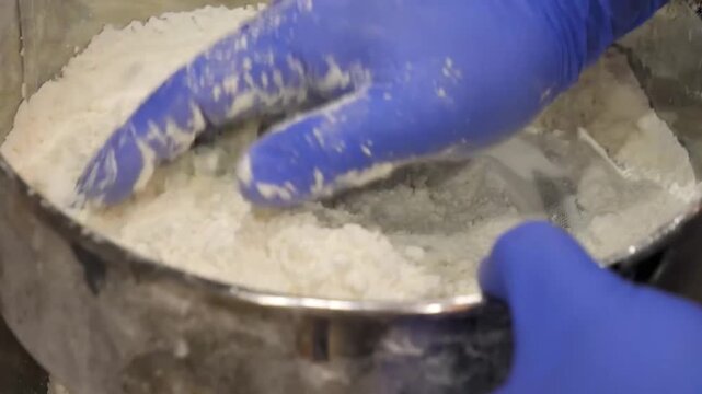 Closeup of a person wearing blue disposable blue gloves using a fine metal sieve to sift white powdery ingredient, likely flour or powdered sugar, into a large metal bowl.