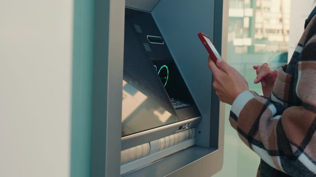 Woman hands hold smartphone, using near field communication technology for contactless cash withdrawal at an outdoor automated teller machine on city street.Cardless cash withdrawal, mobile banking