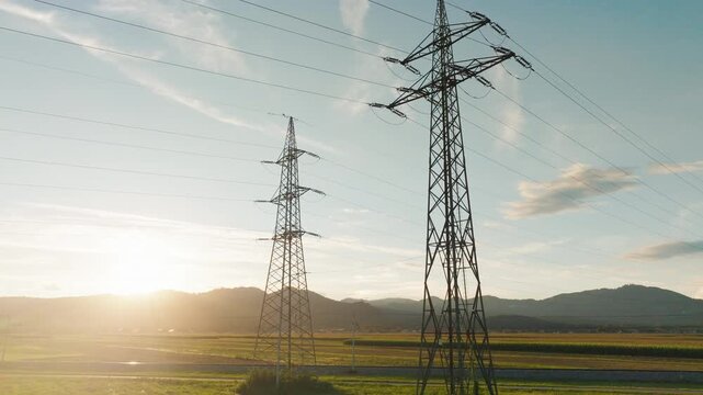 Power lines and towers are visible in a rural area as the sun sets in the background, highlighting energy infrastructure in nature.