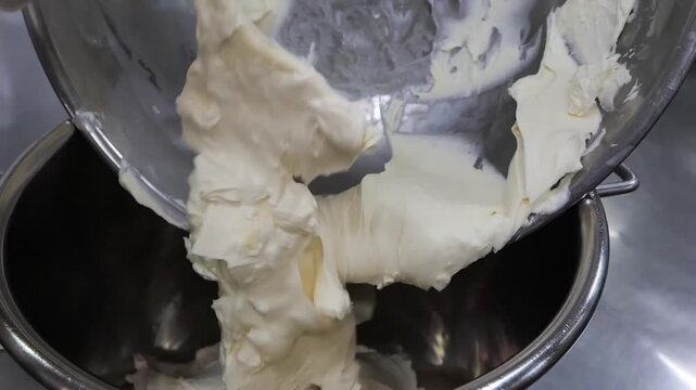 Close-up footage of a large quantity of rich, smooth white buttercream or whipped cream being scooped into a stainless steel mixing bowl. The creamy texture suggests a delicious dessert ingredient, id