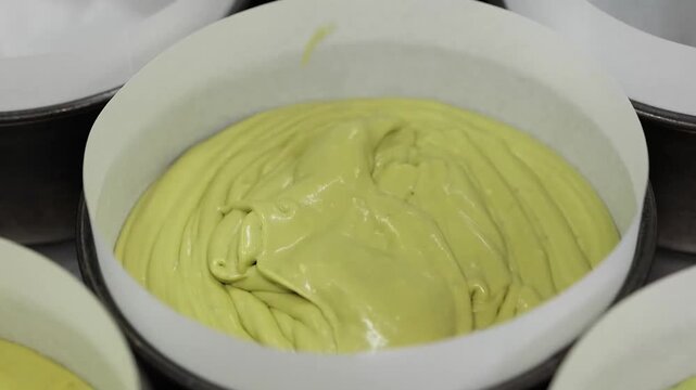 Close-up footage of thick, smooth yellow cake batter or white chocolate ganache being poured into a round metal baking tin lined with white parchment paper. The mixture has a bright, vibrant yellow hu