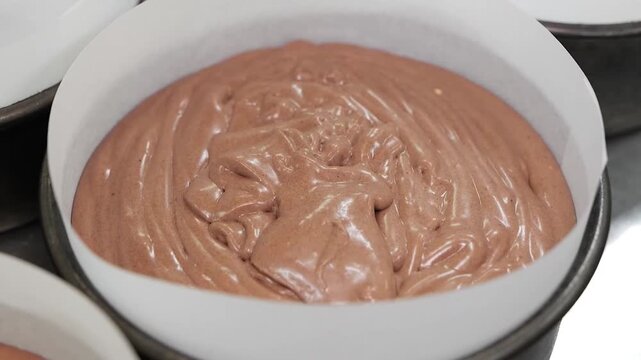 Close-up of thick, glossy chocolate batter being poured into a round baking dish lined with parchment paper. This footage captures the process of homemade baking, dessert preparation, and the rich tex