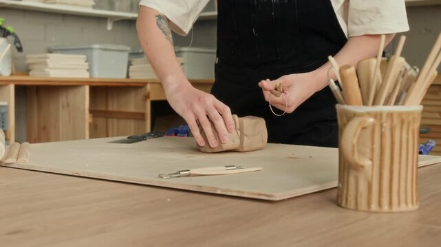 Low angle close up shot of unrecognizable woman in jumpsuit cutting clay with string on wooden table in pottery studio