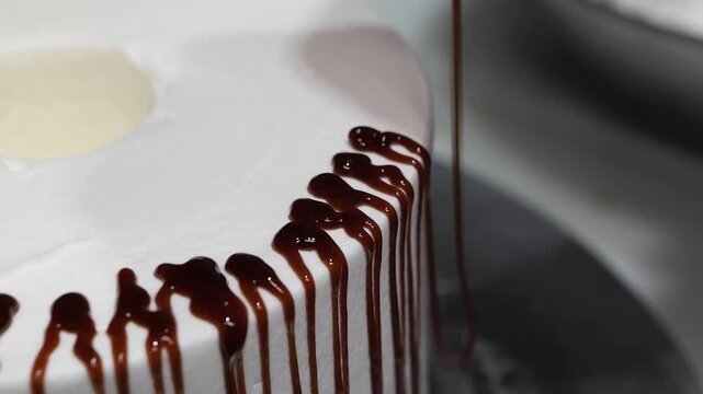 A steady, high-angle close-up shot capturing rich chocolate syrup or ganache being slowly poured onto the edge of a perfectly smooth white buttercream cake, creating a decorative "drip cake" effect.
