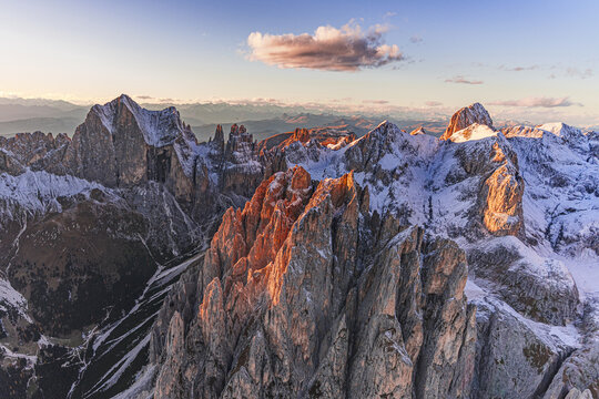 Aerial view of jagged, snow-dusted Dolomites Catinaccio Rosengarten peaks blaze with golden light against the cool blue sky, Trentino-South Tyrol, Italy.