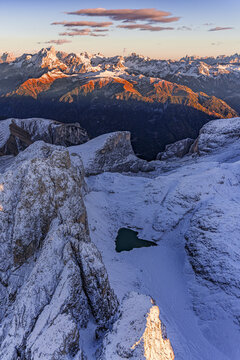 Aerial view of snow-dusted peaks kissed by the sun's fiery glow, serene Antermoia lake nestled amidst rugged, icy terrain, Canazei, Trentino-South Tyrol, Italy.