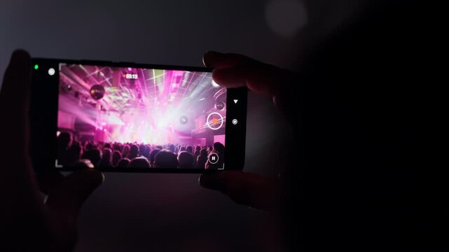 Hands hold a smartphone, recording a vibrant live concert with dazzling stage lights and a cheering crowd. Capturing an immersive event experience from a personal perspective