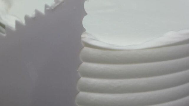 A detailed, close-up shot of a white buttercream cake being decorated using a scalloped cake comb to create smooth, horizontal ridges on the side of the dessert.