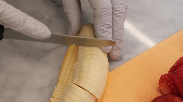 A close-up shot of a person wearing white protective gloves using a stainless steel knife to slice a peeled, ripe banana into small, round pieces on a bright orange cutting board. Fresh red strawberri
