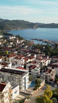 Vertical video of a coastal town by the sea, featuring red-roof houses, a marina, and a calm bay surrounded by green hills. Captured from a drone.