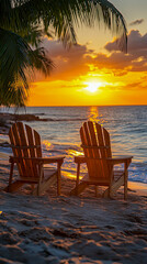 Fototapeta premium Two wooden Adirondack chairs sit on sandy beach foreground facing brilliant orange sunset over ocean, framed by palm fronds