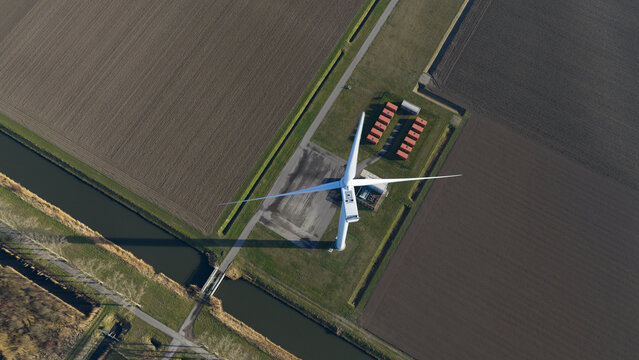 Aerial view of a single wind turbine standing in flat agricultural fields next to a narrow canal.