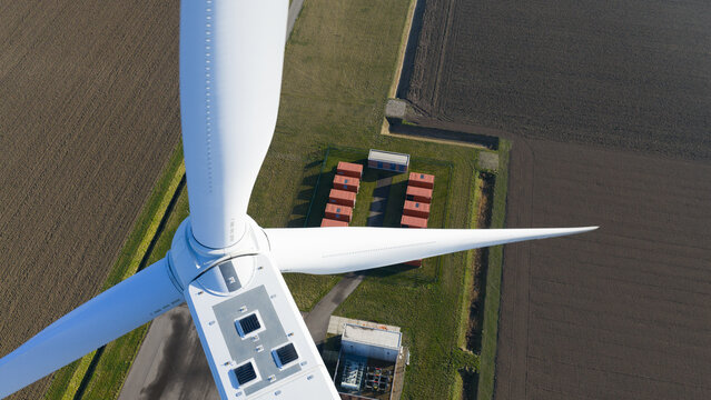 Aerial view of a wind turbine nacelle and white blades above a maintenance area with red containers and agricultural fields.