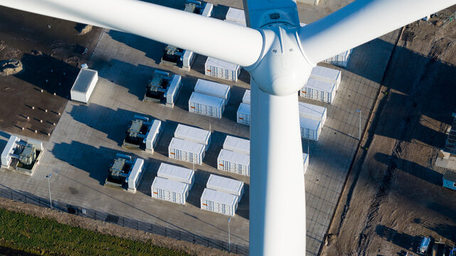 Aerial view of a wind turbine hub and blades overlooking a battery energy storage system with white containers and electrical transformers in Dronten, Flevoland, Netherlands.