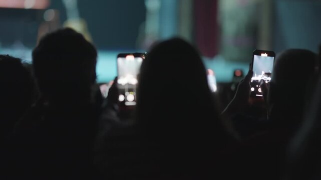 Silhouetted audience at a dark concert or festival records the performance on their smartphones. The bright screens illuminate the crowd's hands, capturing the modern live event experience