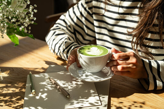 woman drinking matcha latte while sitting at wooden table with notebook and pens, cozy morning and mindfulness concept
