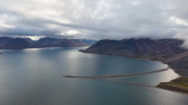 Curved shoreline and sandspit frame Rumilet Bay in Chukotka. Drone footage captures a smooth pan right over water and tundra. Early autumn morning clouds hang low over the mountains