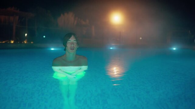 A serene young person relaxes in a vibrant blue-green lit swimming pool at night, eyes closed in contemplation. Steam rises gently around them in this tranquil, atmospheric shot.