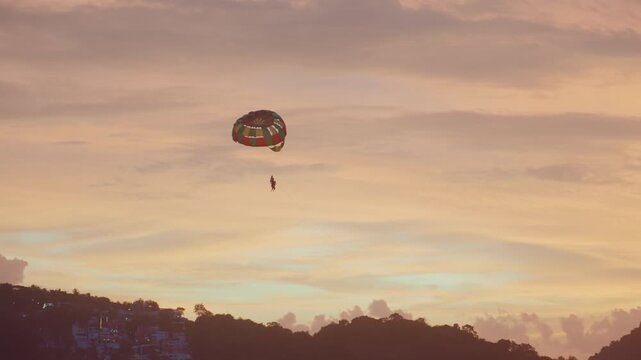 Lone rider floats under colorful canopy at dusk. Relaxed scene of parasailer over tranquil ocean and hills. Serene image of lone parasailer gliding beneath vibrant sunset sky over calm waters