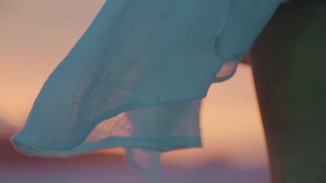 Silken fabric sways with ocean wind during sunset. Barefoot woman moves gracefully along beach during twilight. Relaxed lady in flowing attire enjoys seaside walk with rhythmic steps at sunset