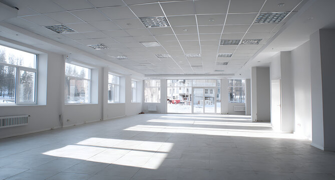 An empty, brightly lit interior space with large windows and a grid ceiling, casting sunlight across the tiled floor