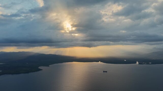 Static high altitude drone captures sunset over Palawan mountains with dramatic crepuscular rays breaking through heavy clouds casting golden light across calm bay water where a lone cargo ship rests